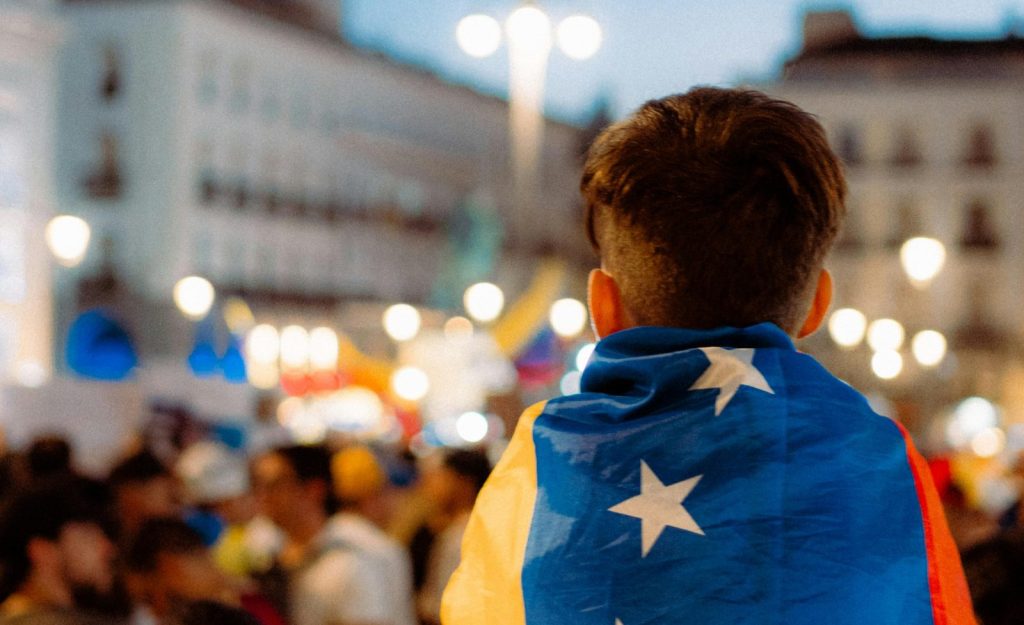 A vibrant city scene with a young man draped in the Venezuelan flag at a nighttime gathering.