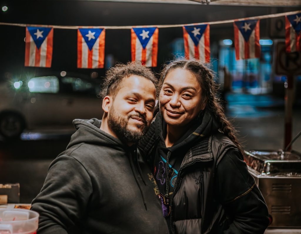 Two smiling people at a Puerto Rican food stall with Puerto Rican flags in Seattle.
