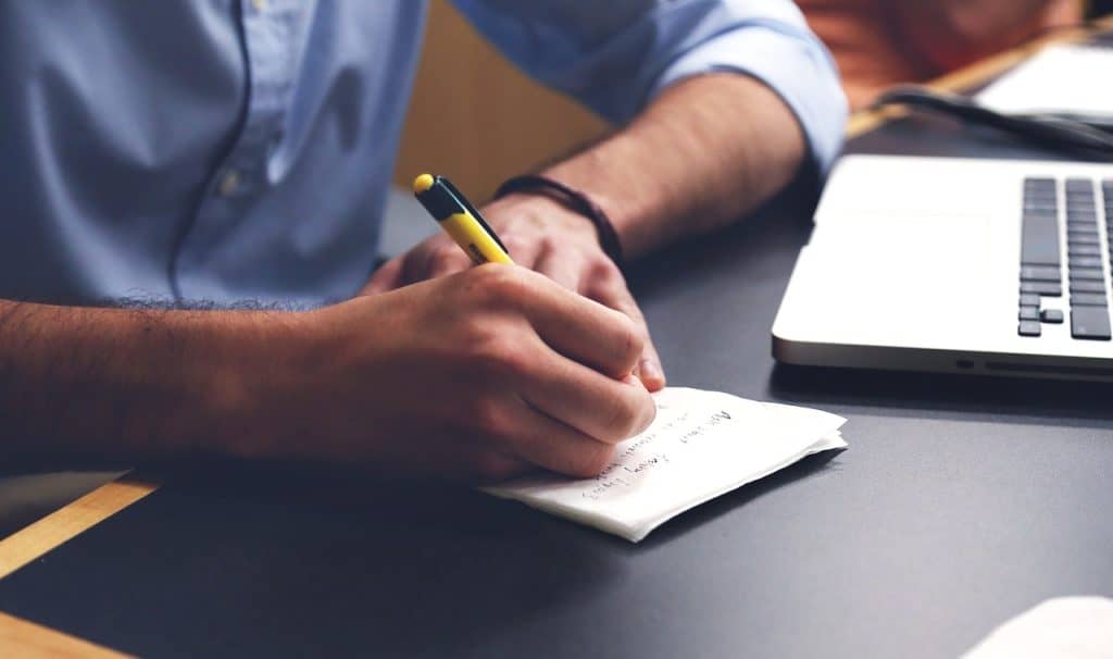 Person taking notes with a yellow pen at a desk with a laptop.