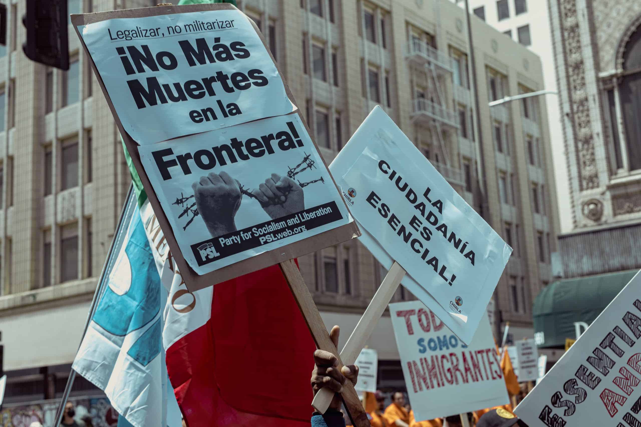 Protesters hold signs against border deaths and militarization in an urban setting.
