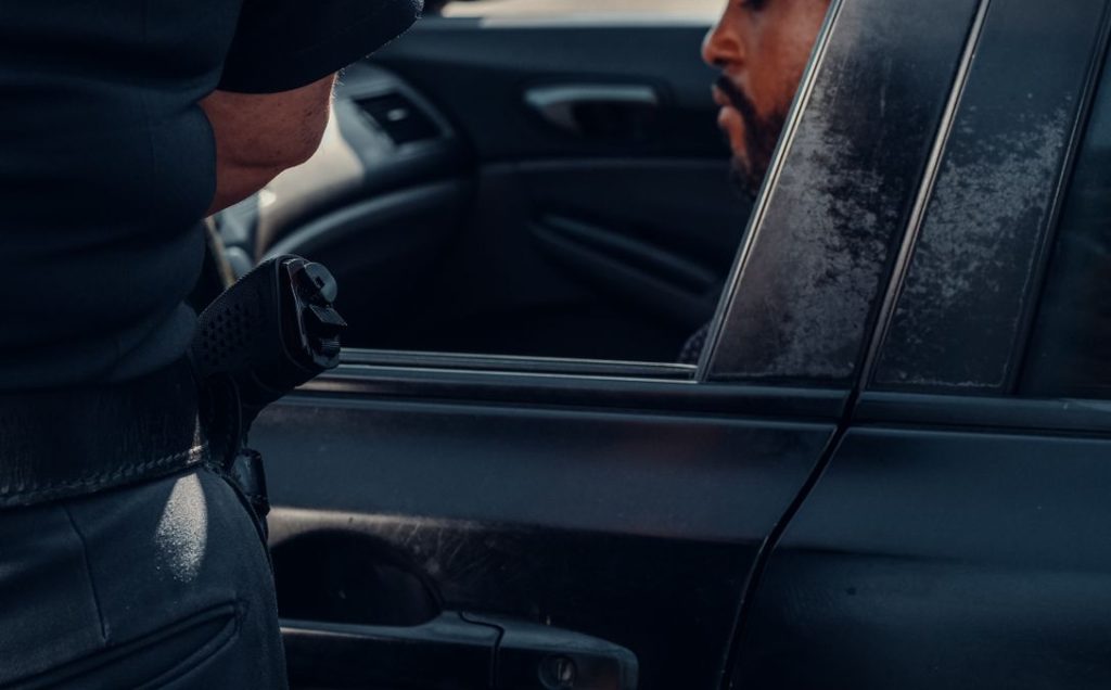 Law enforcement stops a man seated in a car, captured outdoors in daylight.