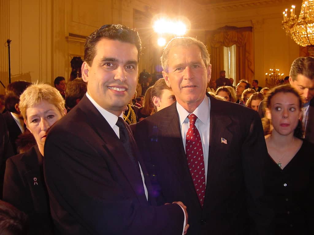 Two men in formal attire at a 9/11 remembrance event with a crowd in the background.